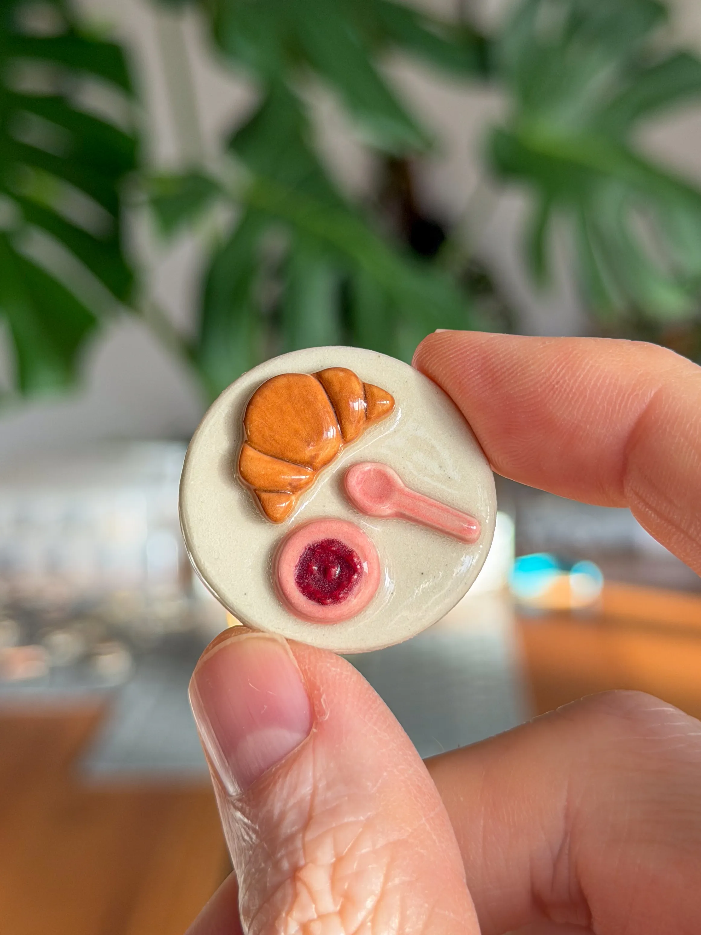Hand-sculpted ceramic magnet of a breakfast plate with a croissant, pink spoon and pot of jam