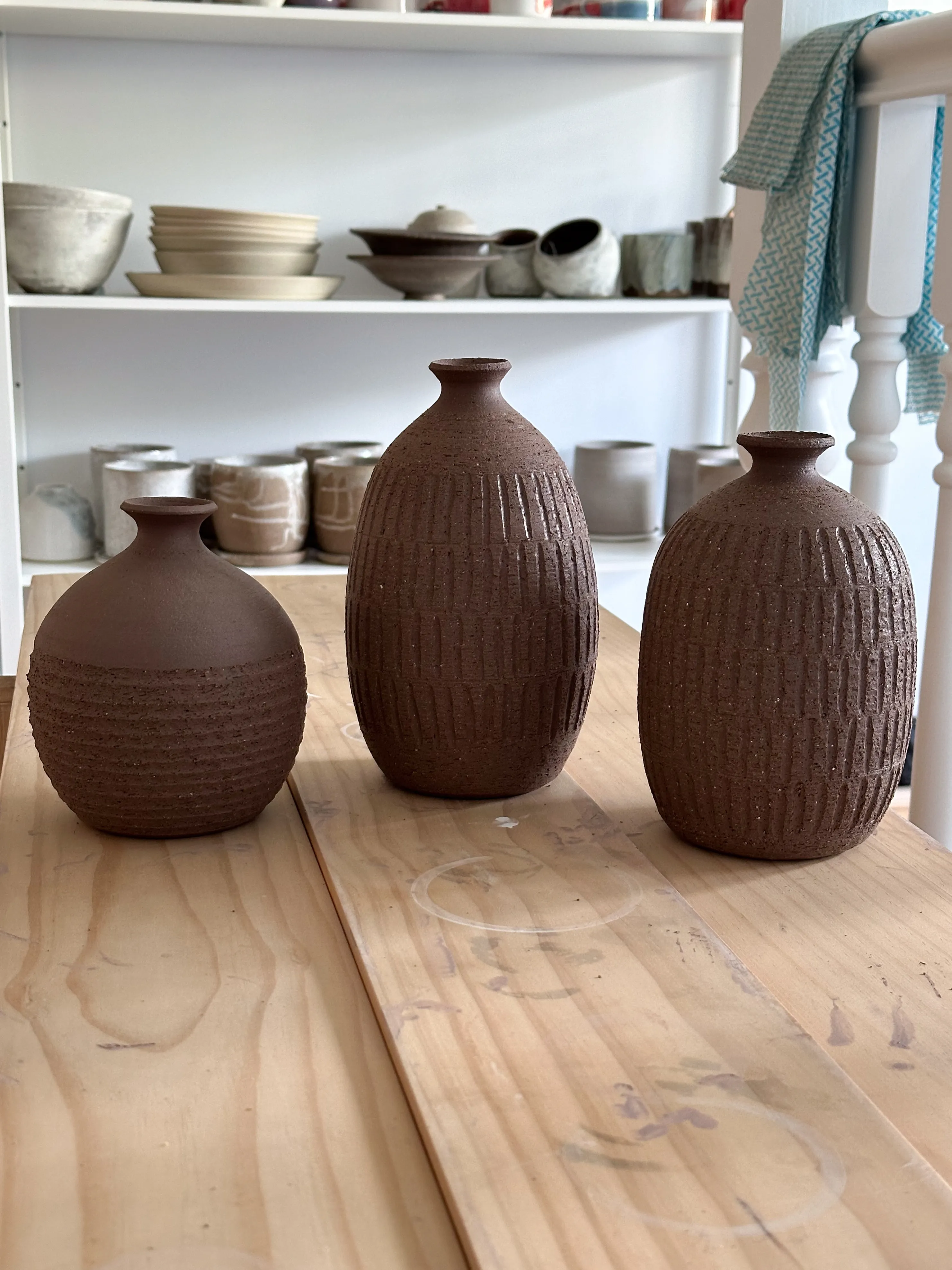 Three dark stoneware bottle vases with carved fork-cut texture, unglazed, on a wooden bench