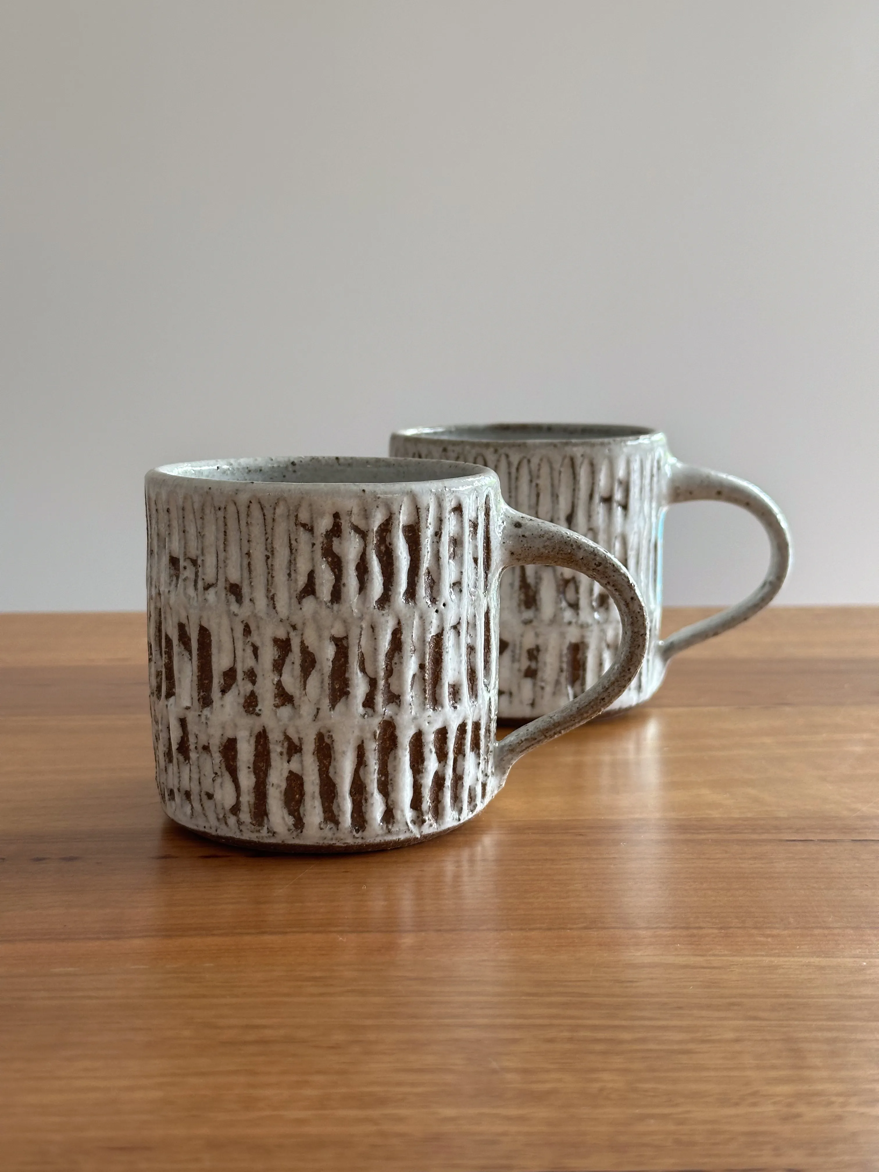 Two wheel-thrown stoneware mugs with vertical fork-cut texture and white glaze pooling