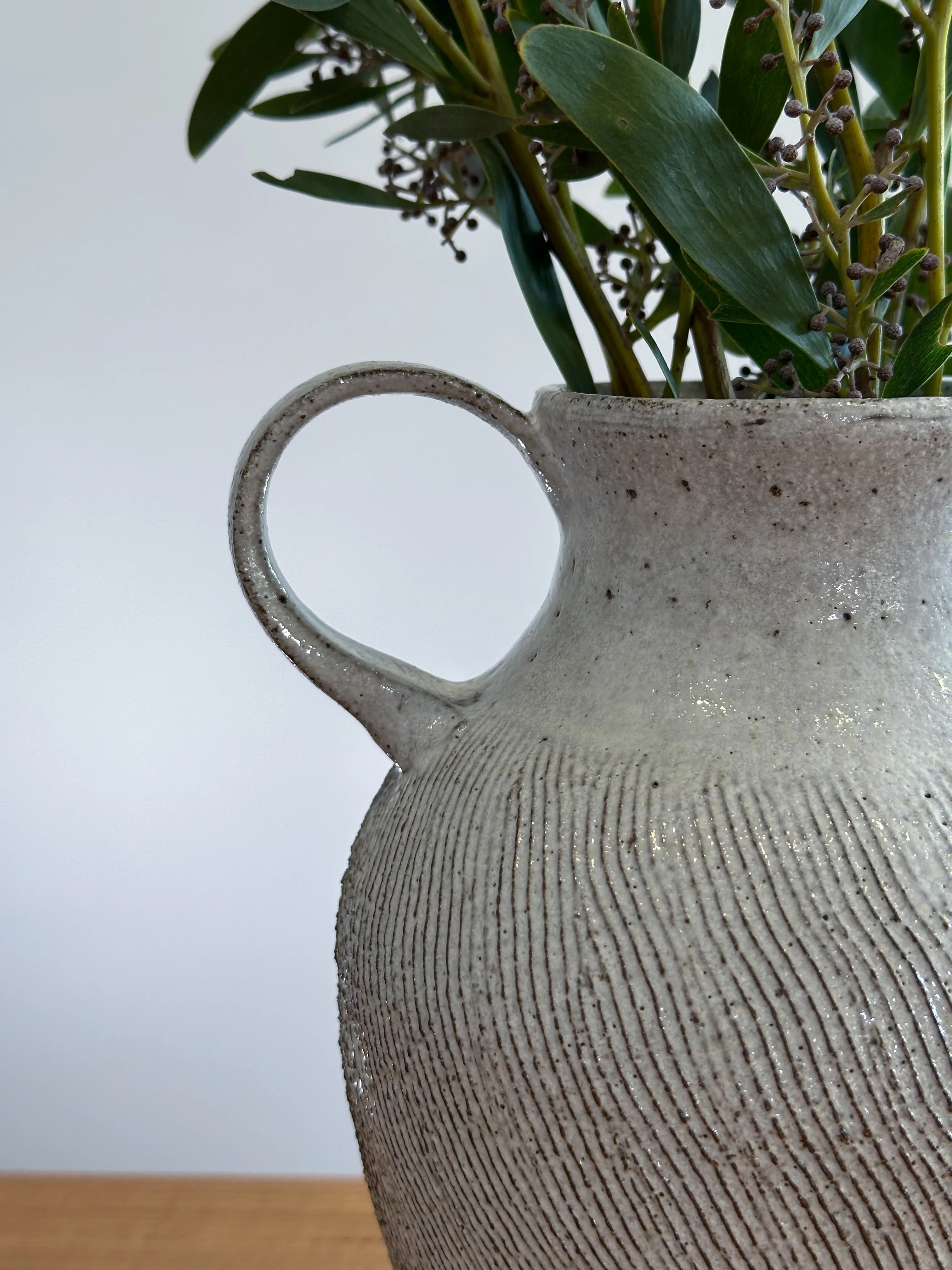 Close-up of a two-handled stoneware vase with vertical wheel-rib texture, holding eucalyptus